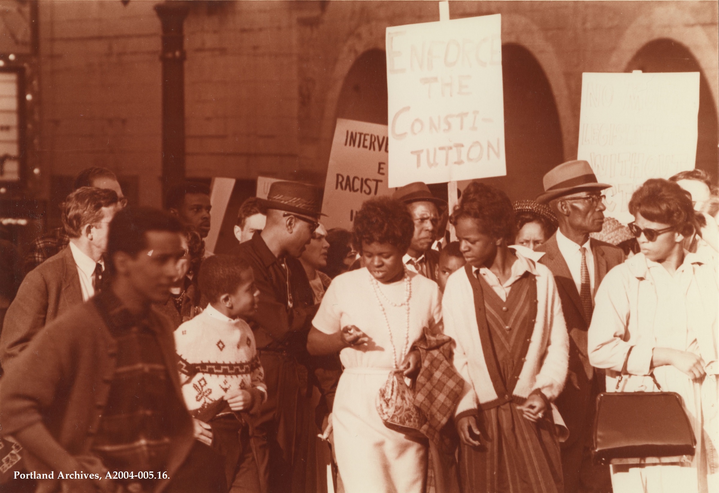 Black and white photograph of people walking in protest with signs during a Civil Rights march. One side reads “Enforce the Constitution.” 1965
